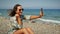 Happy smiling surfer girl taking selfies on smartphone while sitting on beach at sunny windy day. Carefree young brunette woman ma