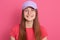 Happy smiling woman looking playful at camera while posing isolated over pink background, wearing red t shirt and baseball cap,