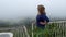 Happy smiling woman standing at green abyss with thick fog on a view point with hammock net high in mountains on edge