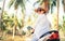 Happy smiling woman in straw hat and sunglasses riding motorbike under palm tree.. Careless vacation time concept image