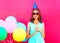 Happy smiling young woman in a birthday cap with an air colorful balloons and lollipop on stick over pink background