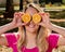 Happy and smiling young woman playing with an orange cut in half and in front of her eyes. Photo in outdoor park and under trees