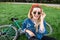 Happy, stylish girl sitting on a green grass in a park near a bicycle, looking into the camera and smiling. Attractive smiling