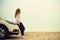 Happy stylish young woman traveler on beach road sitting on white crossover car, holding hat in hand. Banner. Travel, summer