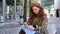 Happy teen redhead girl sitting on big city urban street using smartphone.