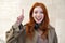 Happy teen redhead girl standing on urban wall background pointing up.