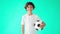 Happy teenage boy in white t-shirt holding soccer ball in one hand and laughing at camera while standing against green