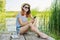 Happy teenage girl in hat with smartphone on wooden pier of lake with reeds