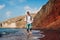 Happy tourist running on Red beach by sea on Santorini island barefooted. Man with backpack traveling