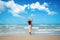 Happy traveler woman in sunhat and dress standing with her arms raised looking tropical beach on summer day. Holiday and summer