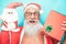 Happy trendy santa claus holding gift and christmas puppet - Fashion senior man having fun wearing xmas costume