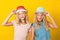 Happy twins sisters wearing summer hats, over yellow background. Summer fashion. Stylish girls with blonde wavy hair, beauty eyes