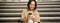 Happy urban girl drinks her takeaway coffee and scrolls feed on smartphone. Asian woman sits on stairs with tea and