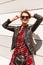 Happy urban young woman in trendy youth clothes with black handbag straightens stylish sunglasses and hair near modern wall on