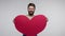Happy Valentine`s day! Handsome cheerful bearded man holding large paper heart, looking at camera with joyful smile, demonstrating
