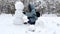 Happy woman building snowman at winter forest. Young girl having active leisure at snowy park. Cheerful female having