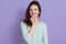 Happy woman with dark hair keeping fist under chin, looks at camera, wears casual attire, poses against lilac background,