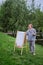 Happy woman draws on the easel with a brush and paints. Woman artist draws nature and trees on paper by the water on the river