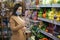 Happy woman in face mask hold phone and choose vegetables in supermarket. Beautiful young business woman choosing