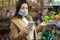 Happy woman in face mask hold phone and choose vegetables in supermarket. Beautiful young business woman choosing