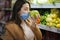 Happy woman in protective mask taking fresh vegetables while standing by groceries in supermarket. Beautiful young girl