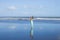 Happy woman walking barefoot on empty beach. Full body portrait. Slim Caucasian woman wearing long dress. Water reflection. Blue