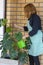 A happy woman waters a large monstera flower in a room with beige brick walls with a green watering can. A girl in an apron is