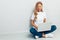 Happy woman wearing white t-shirt sitting on the floor with laptop on grey background