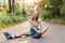 Happy young adult woman in rollerblades, sitting on road, wearing beige t shirt and jeans short, looking directly at camera and