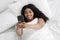 Happy young african american lady lying in bed and having video call on smartphone, lying in bed under white blanket