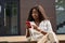 Happy young African woman model using smartphone sitting outdoors.