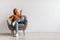 Happy young Asian woman with remote relaxing under air conditioner, sitting in armchair against white wall, mockup