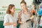 Happy young assistant of gardening shop in apron and shirt showing small shovels to female customer