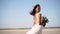 Happy young bride with bouquet of fresh flowers going at desert landscape. Elegance lady in gorgeous wedding dress walks