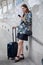 happy young business woman standing with hand luggage using mobile phone . traveling girl with suitcase and  smartphone in city