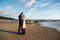 Happy young couple hugging and kissing on the beach. Cold autumn weather, trees in the background.