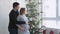 Happy young family. Husband hugs him pregnant wife near beautiful decorated Christmas tree look at window
