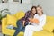 Happy young female lesbian couple in casual clothes sitting together on yellow sofa at home, are reading novel book