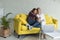 Happy young female lesbian couple in casual clothes sitting together on yellow sofa at home, are watching funny movie on