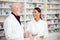Happy young female and senior male pharmacists standing in front of shelves with medications and talking