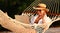 Happy young female wearing straw hat waving while having video call on digital laptop, relaxing in the hammock on tropical beach