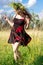 Happy young girl in short dress with a colorful garlang made of wild flowers on her head dances and laughs on the meadow.