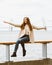 Happy young girl sitting on waterfront at pier in the port, enjoying life and waving his arms. Woman with long hair smiles and