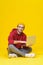 Happy young hipster man in red hoodie sitting on the floor holding laptop studying or working. Smart freelancer