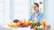 Happy young housewife sitting in the kitchen preparing food from a pile of diverse fresh organic fruits and vegetables