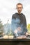 A happy young man cooking meat burgers on barbecue grill - leisure, food, people and holidays concept