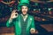 Happy young man in green suit stand in pub and hold golden coins. He look at one of them and amaze. Guy wear St. Patrick