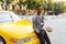 Happy young man a mixed race holding in hand a cup coffee, seated on a hood yellow car,  on a street background.