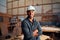 Happy young man in safety helmet looking at camera with arms crossed in woodworking factory