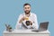 Happy young man sitting at desk in office, holding money terminal and credit card, blue background